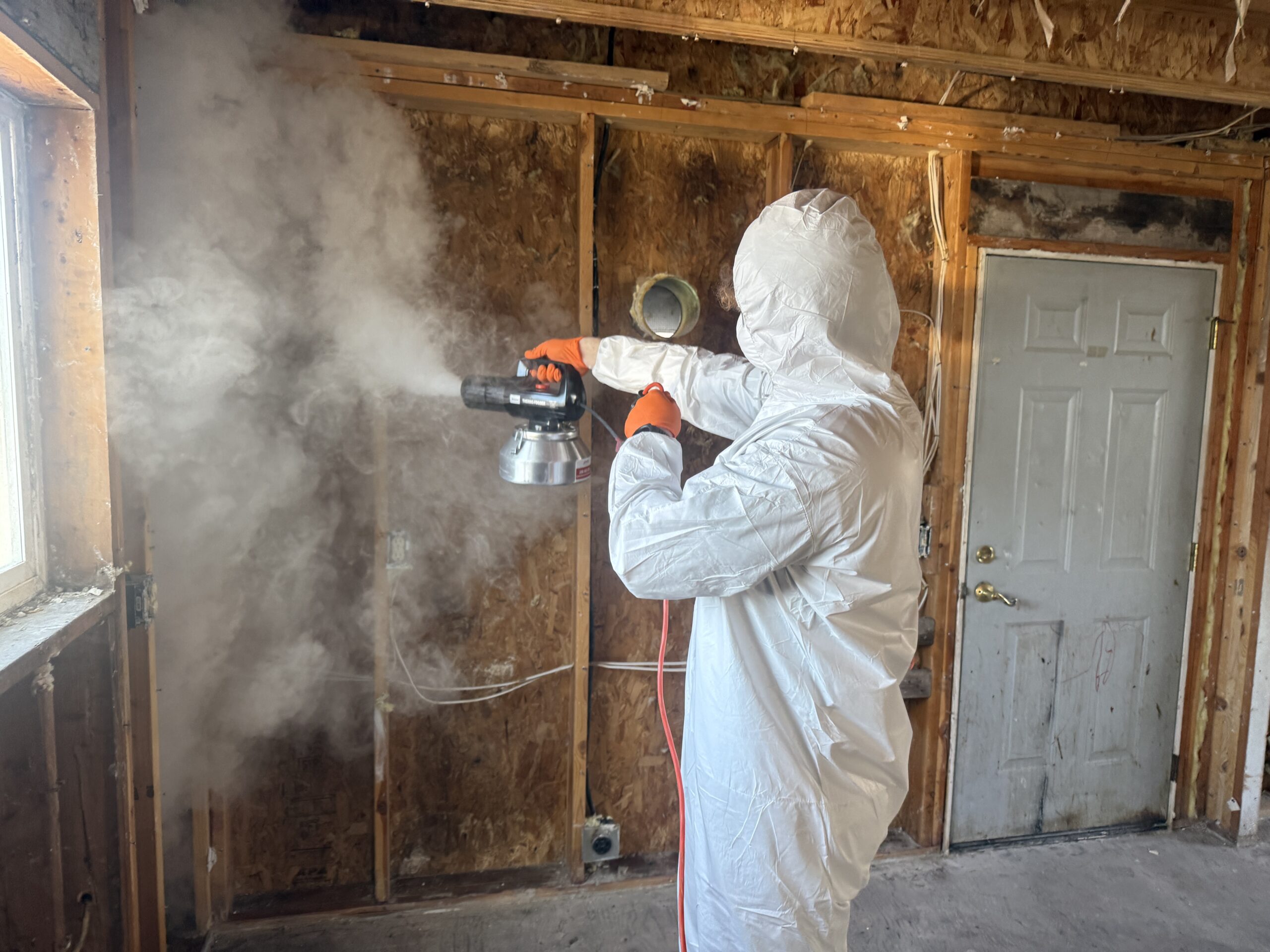Technician in full protective equipment applying treatment to a room stripped to the studs