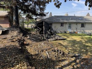 Collapsed, burned shed and charred debris in a backyard, with a scorched pine tree and a brick house behind