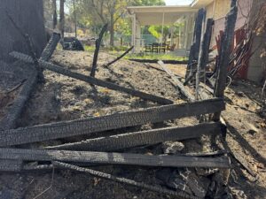 Charred fence boards and burned posts lying on scorched ground, with a home's covered patio in the background