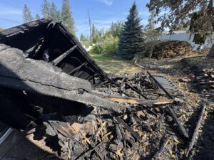 Collapsed shed roof with charred rafters and a pile of burned lumber and debris in a grassy yard