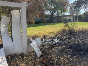 White vinyl fence panels warped and melted by heat next to a burned strip of yard, with a ranch house across the lawn