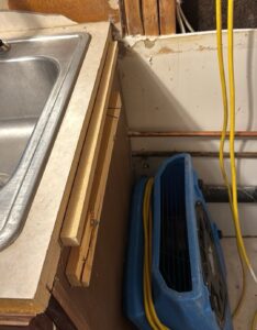 Blue air mover aimed at a cut-open wall beside a kitchen sink cabinet, with copper water lines exposed