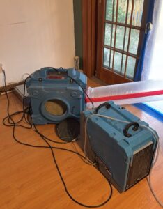 Blue air scrubber and dehumidifier set up on a wood-look floor beside a patio door sealed off with plastic sheeting