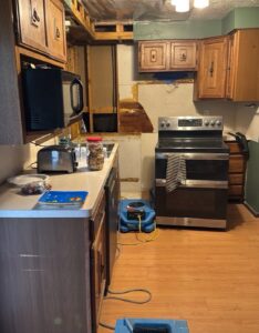 Kitchen with the lower drywall cut out and an upper cabinet removed to expose wall framing, with a blue air mover drying the floor in front of the range
