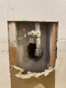 Square section of drywall cut away to expose a galvanized water pipe and red-handled shutoff valve inside the wall
